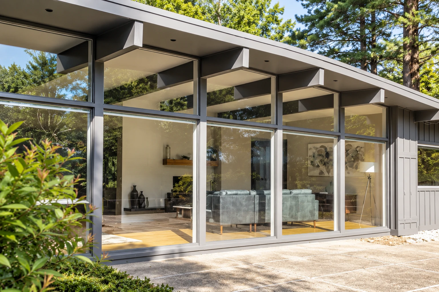 Modern all-season room with broad glass walls and dark framing, showing a furnished interior and wooded surroundings.