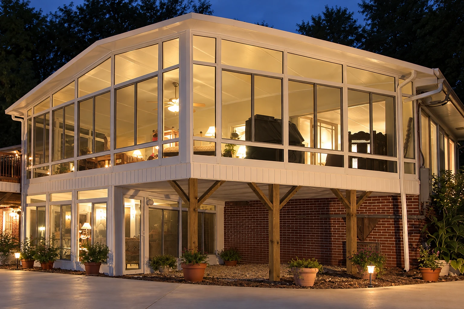 Two-story custom sunroom with white-framed glass walls, warmly lit interiors and timber supports, shown at dusk.