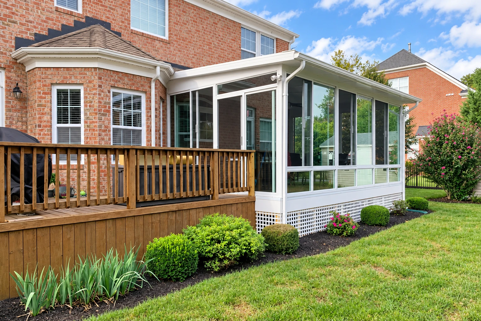 White-framed sunroom addition beside a wooden deck on a brick house, with large windows and garden beds.