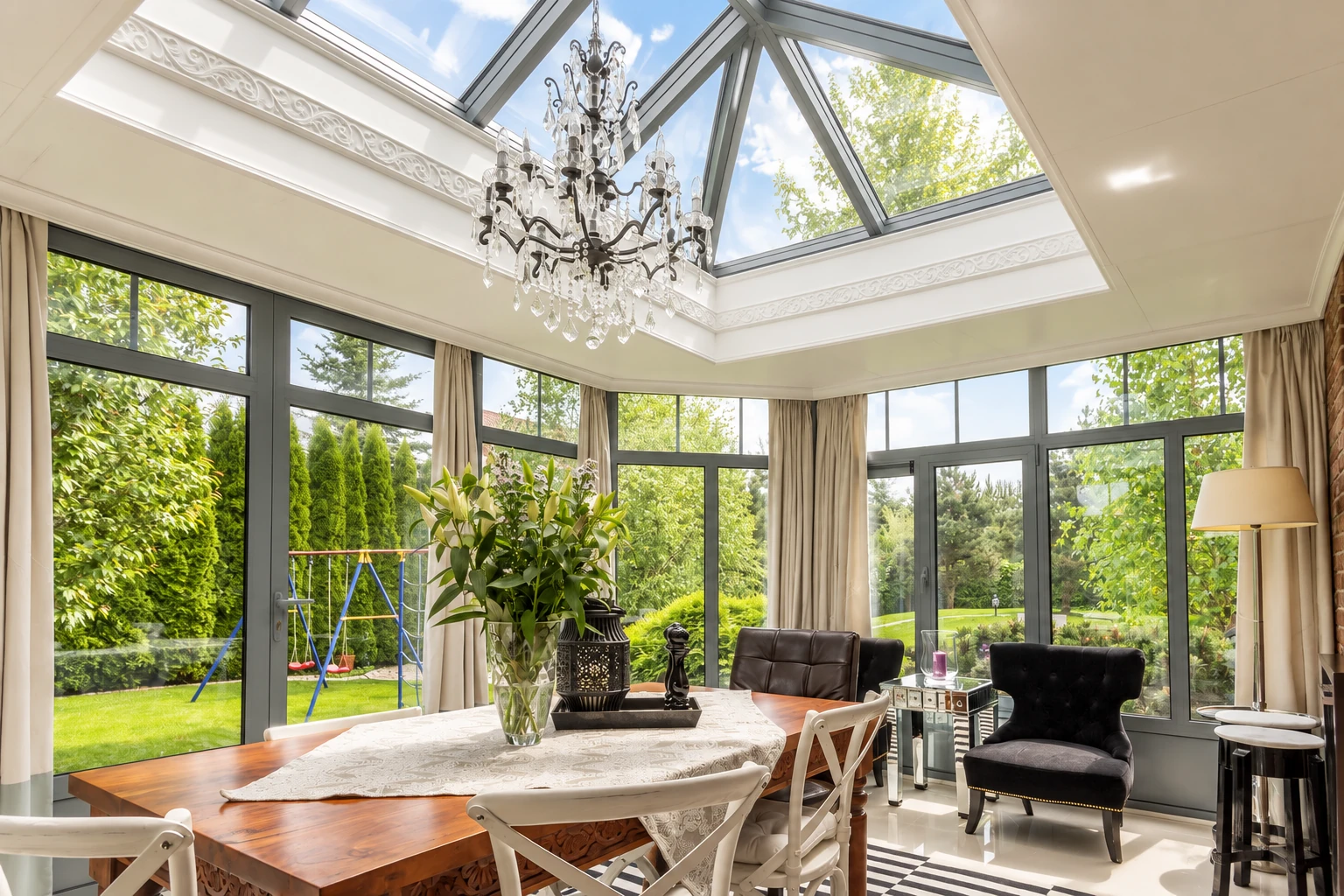 Three-season sunroom with a glass-paneled vaulted ceiling, chandelier, dining table and garden views.